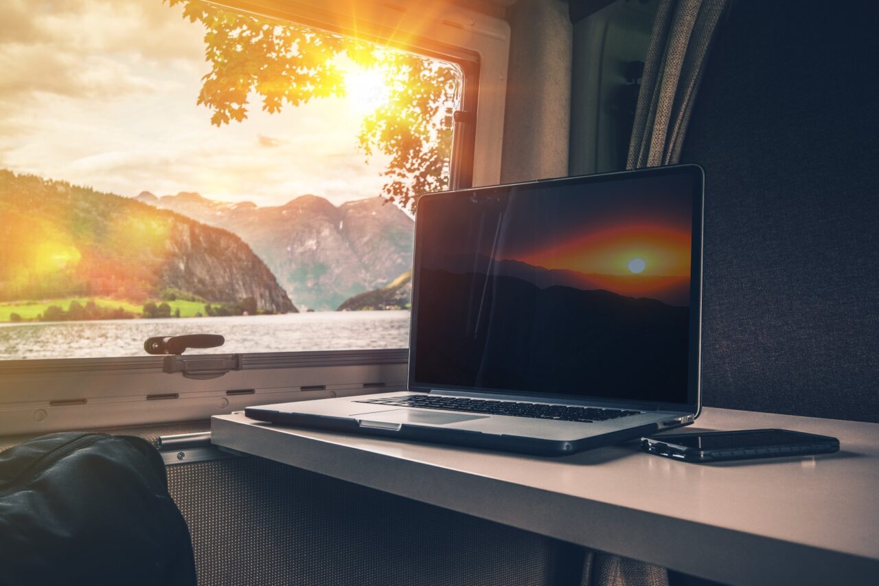 Ein Laptop und Smartphone liegen auf dem Tisch in einem Campingfahrzeug. Durch das Fenster sieht man einen See, Berge und den Sonnenuntergang.